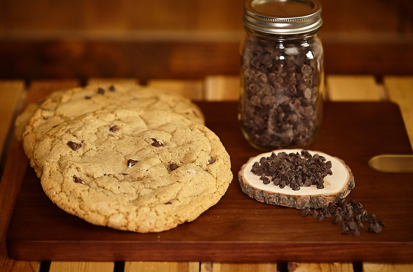 three freshly baked chocolate chip cookies on a cutting board with chocolate chips