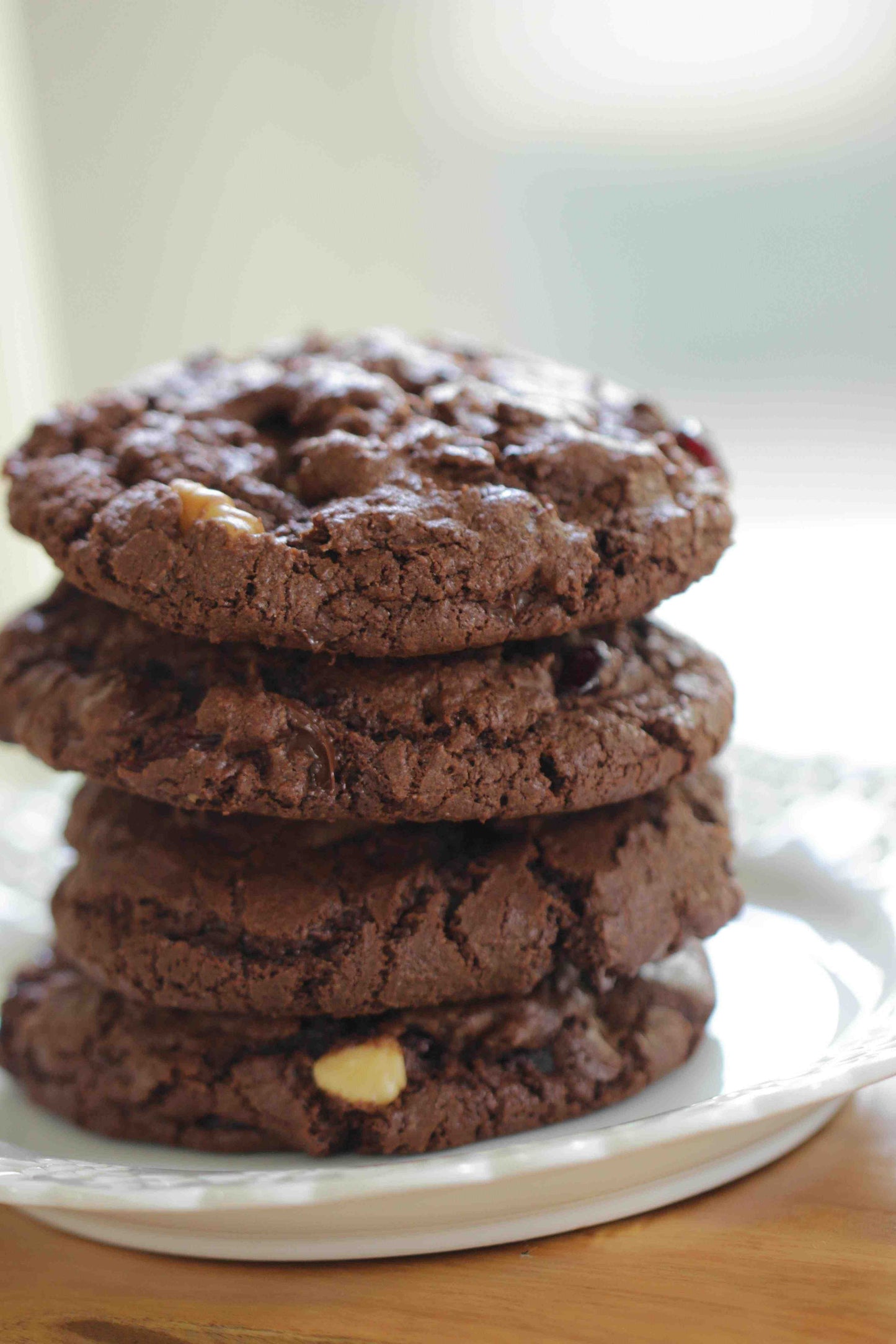 A stack of three double chocolate chip cookies on a white plate, with a blurred background.
