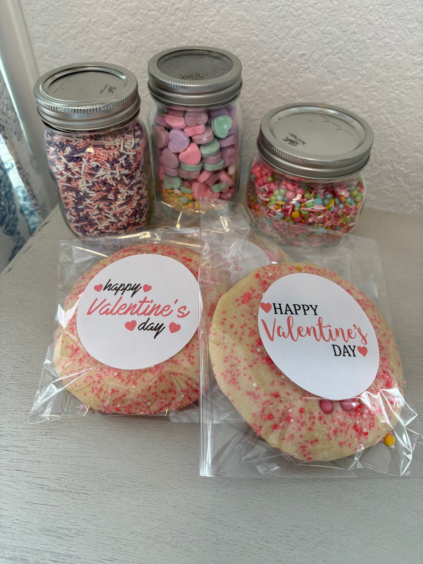 Packaged sugar cookies with 'Happy Valentine's Day' and colorful sprinkles on top, displayed alongside jars of decorative sprinkles.