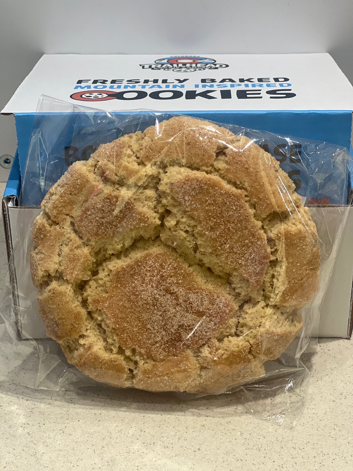 Large cinnamon sugar cookie in front of a box labeled 'Freshly Baked Mountain-Inspired Cookies'.