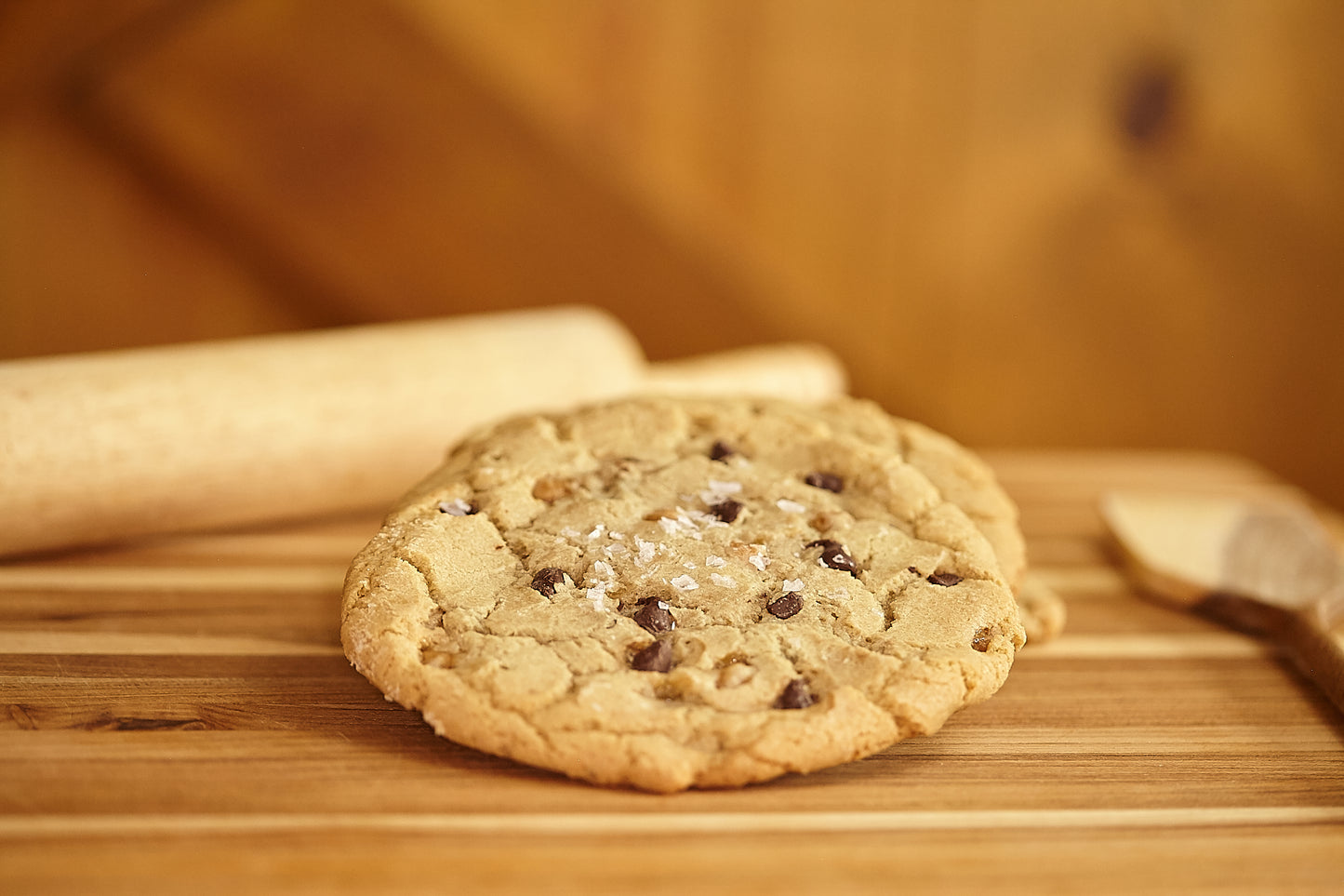 A cookie with chocolate chips and a sprinkle of sea salt, placed on a wooden surface with a rolling pin and a spoon in the background, suggestive of a baking or cooking scene.