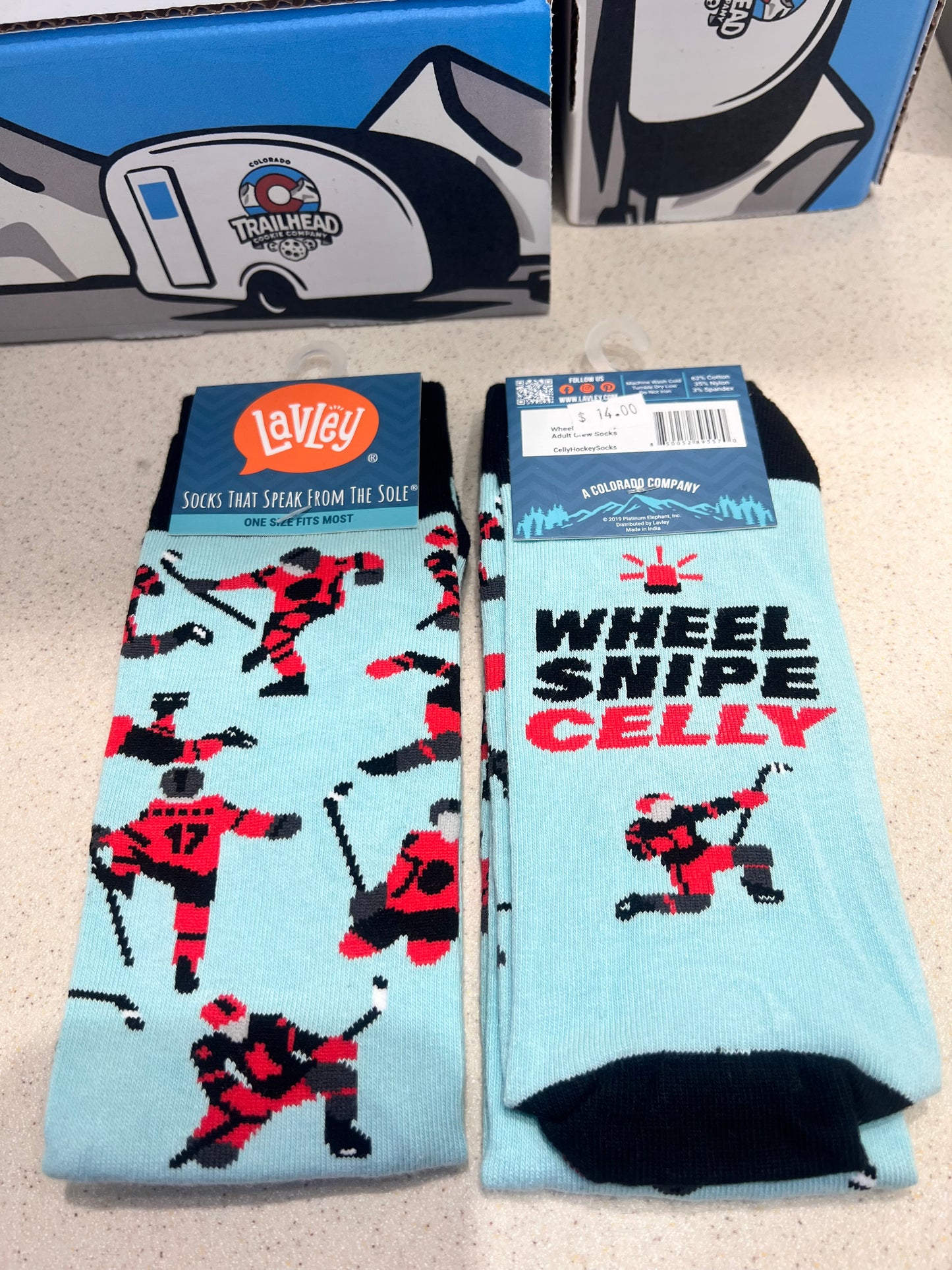 Light blue socks with red and black graphics and 'Wheel Snipe Celly' hockey text referring to celebrating on a beige surface.