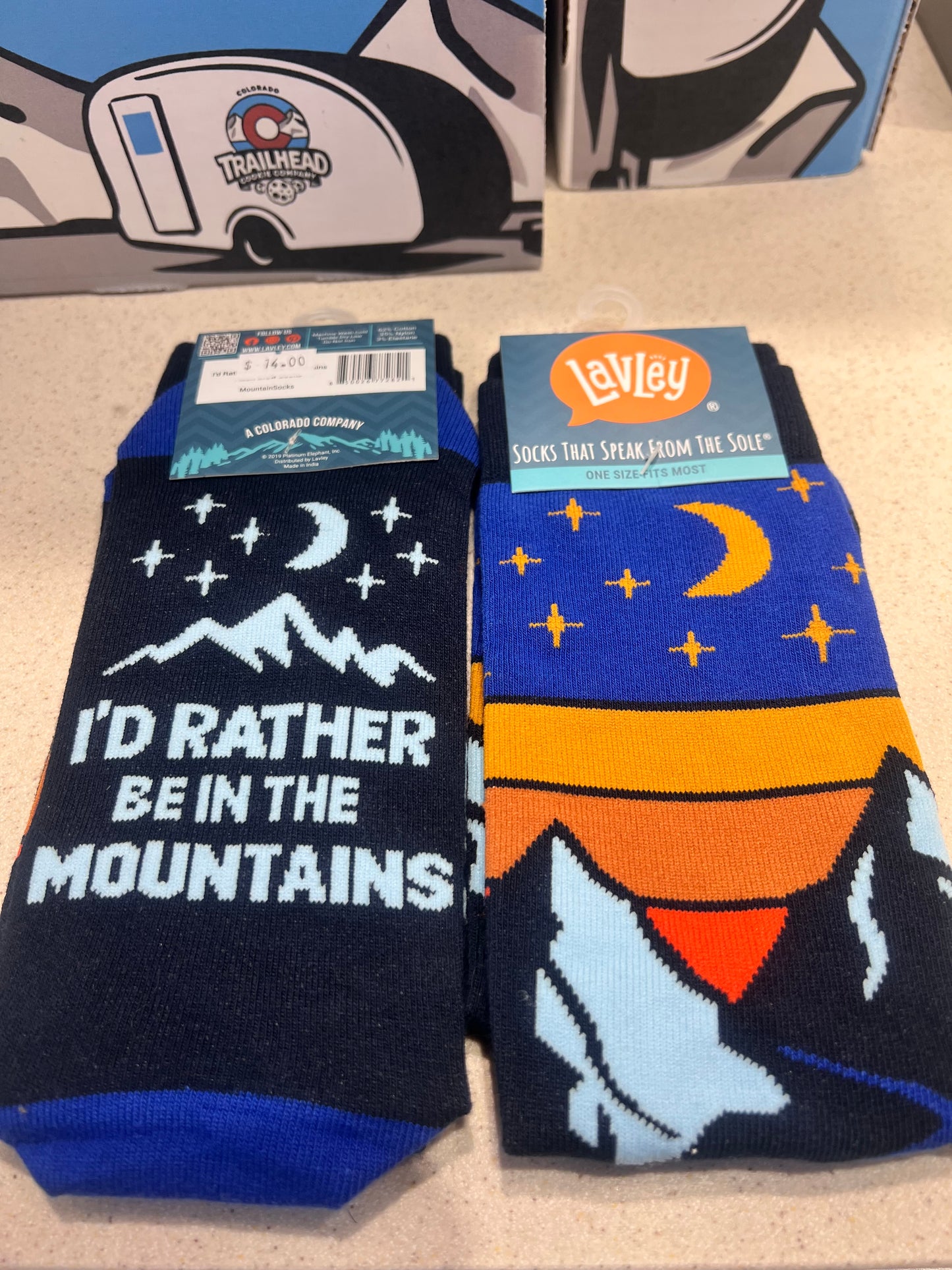 Colorful socks with mountain design and "I's rather be in the mountains" branding on a beige surface