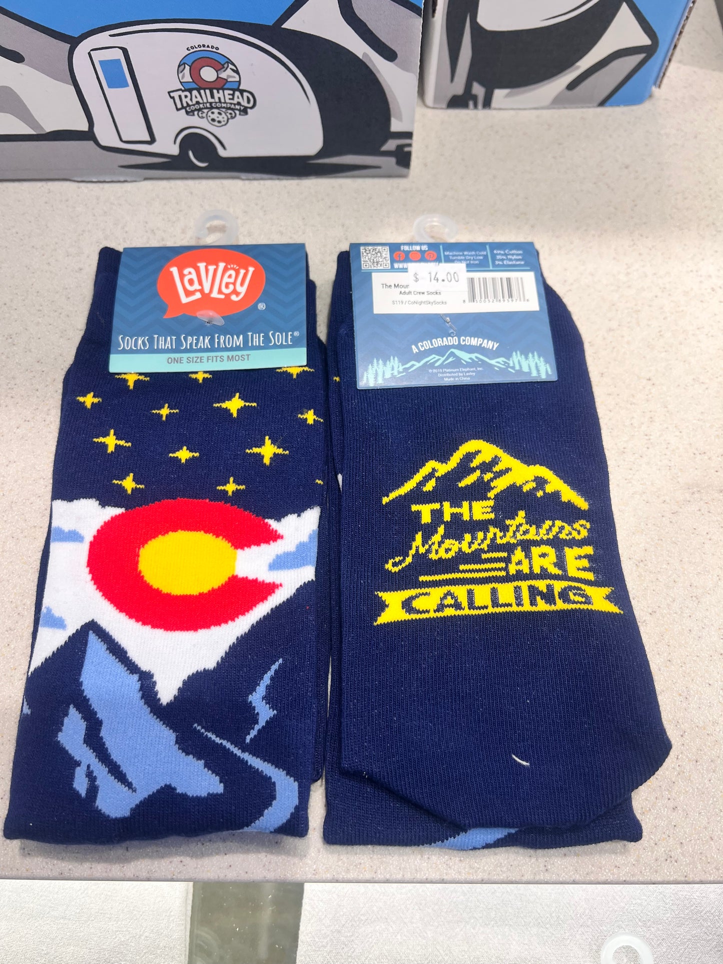 Pair of blue socks with mountain and Colorado state logo design on a gray surface with the text  "the mountains are calling"