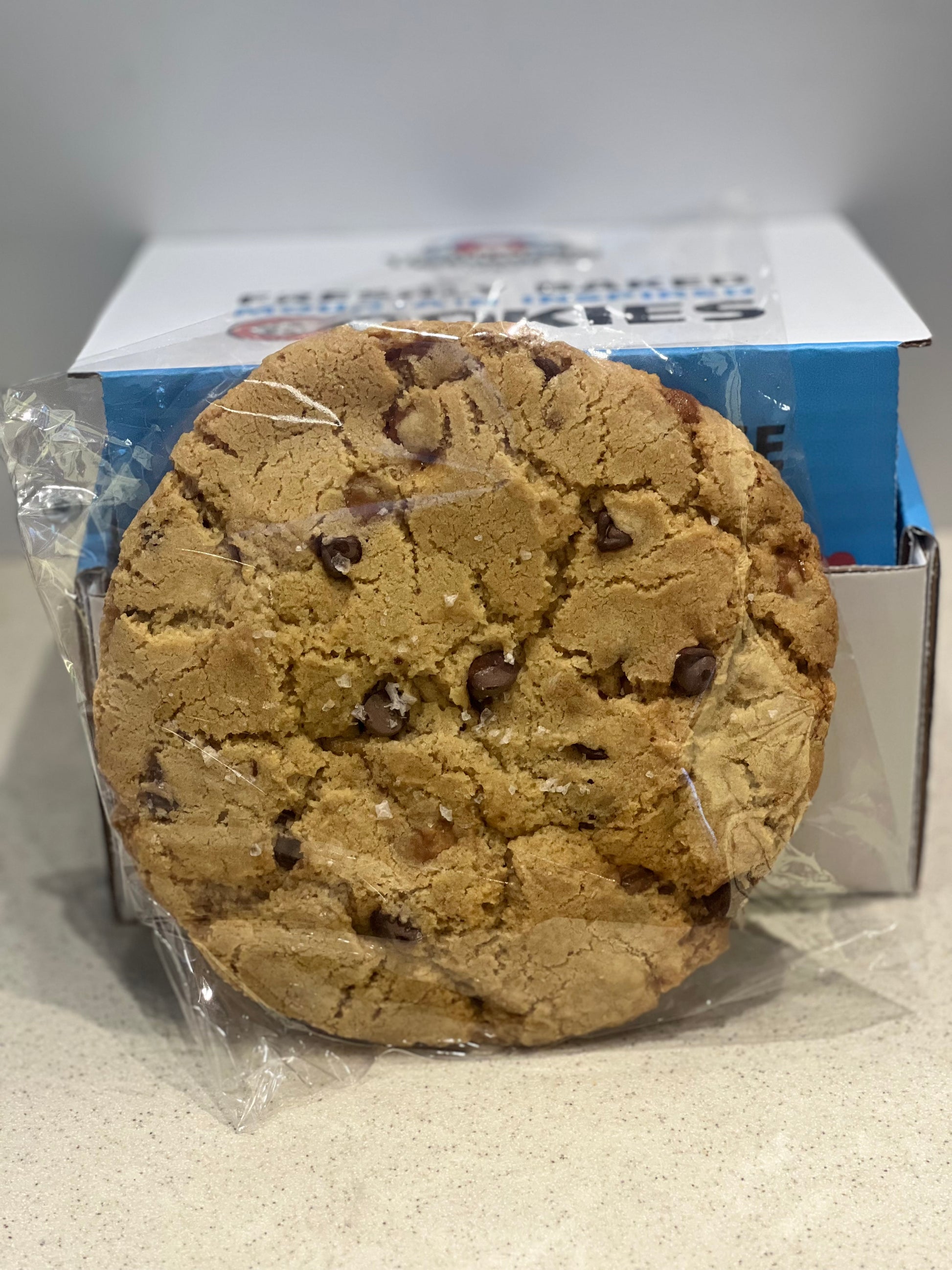 Chocolate chip cookie in clear plastic wrap with a blue box in the background